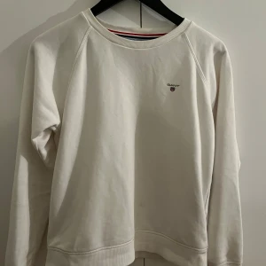 Vit sweatshirt från GANT - Vit sweatshirt från GANT. Tröjan är i ett bra skick men har en liten fläck på framsidan, kanske kan gå bort med rätt medel. Tröjan är i St 15 år, ungefär Xs/S. Tvättas innan den säljs. 