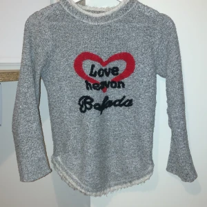 Grå tröja med hjärtmotiv från MIN'S - Mysig grå tröja från MIN'S med ett rött hjärta och texten 'Love Never Fades' på framsidan. Tröjan har långa ärmar och en fluffig kant vid halsen och nederkanten. Perfekt för kyliga dagar.Tröjan är jättebekväm och varm och passar ungefär en xs då jag inte vet storleken(10A)