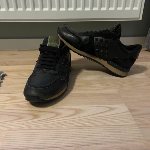 Valentino Rockrunner - Snygga svarta sneakers från Valentino med ikoniska nitar på sidorna och hälen. Skorna har en kombination av skinn och mocka, vilket ger en lyxig känsla. Perfekta för en stilren look. Självklart äkta, alla nitar sitter kvar däremot har det slitits på någon. En söm har lossnat (se bild 5) Även lite slitet inuti skon vid baksidan av hålen, men inget man märker av när man använder dem. Endast skorna följer med. Perfekta till vintern! Skriv till mig vid frågor eller för fler bilder. 