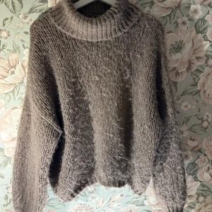 Brun stickad tröja - Mysig brun stickad tröja med en hög krage och långa ärmar. Perfekt för kyliga dagar och ger en avslappnad look. Tröjan har en fluffig textur som gör den extra bekväm.