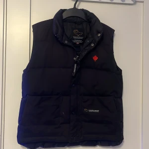 Svart dunväst från Canada Goose - Säljer en svart dunväst från Canada Goose med en broderad röd lönnlöv på bröstet. Västen har en dragkedja och knappar framtill för extra skydd mot kyla. Perfekt för kyliga dagar.