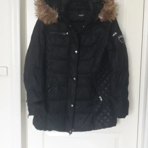 Svart dunjacka från Rockandblue - Snygg svart dunjacka från Rockandblue med äkta päls på huvan. Jackan har dragkedja och knappar framtill samt flera praktiska fickor. Perfekt för kyliga dagar. Använt en månad.