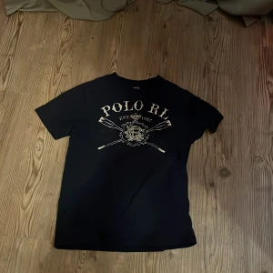 Svart t-shirt från Polo Ralph Lauren - Snygg svart t-shirt från Polo Ralph Lauren med ett vintageinspirerat tryck i guld på framsidan. Perfekt för en avslappnad stil med en touch av elegans. Tillverkad i mjukt material för extra komfort.