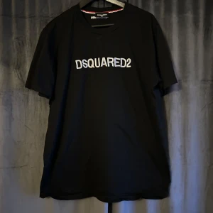 Svart t-shirt från DSQUARED2 - Snygg svart t-shirt från DSQUARED2 med vit logga på bröstet. Tröjan är i bra skick men används ej längre. Storleken är XXL men skulle säga att den passar som en L-XL. Nypris: 2500kr. Kontakta gärna vid frågor ☺️
