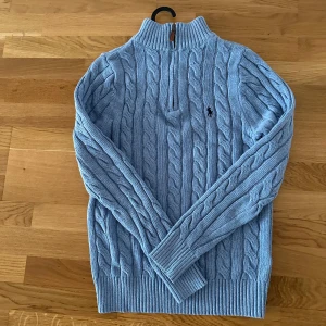 Ljusblå stickad tröja från Polo Ralph Lauren - Säljer en stilren ljusblå stickad tröja från Polo Ralph Lauren med kabelstickat mönster och en liten broderad logga på bröstet. Tröjan har en dragkedja vid halsen och ribbade muddar vid ärmslut och nederkant.