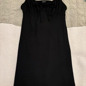 Svart klänning från Gina Tricot - Snygg svart klänning från Gina Tricot med smala axelband och knytning framtill. Perfekt för en stilren look. Klänningen har en enkel och elegant design som passar till många tillfällen.