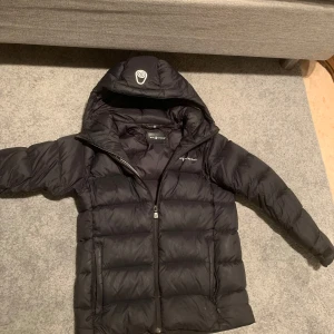 Svart dunjacka från Sail racing  - Säljer en svart dunjacka från Sail racing med huva och dragkedja. Jackan är quiltad och perfekt för kalla dagar. Den har en stilren design och är långärmad. Varan har tecken på användning där sömmarna har gått upp men går att reparera hos din lokala skräddare. Nypris ca 2 500kr. Storlek: 170cm