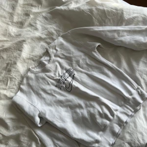 Vit sweatshirt från Nike - Säljer en stilren vit sweatshirt från Nike med deras ikoniska logga broderad på framsidan. Tröjan har långa ärmar och en klassisk rund halsringning. Perfekt för en avslappnad look. Köpte på Plick för några år sen har ej kommit till användning. 
