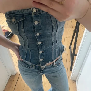 Jeans korsettopp - Snygg jeans korsettopp med knappar framtill och smockdetalj baktill. Toppen är axelbandslös och har en croppad passform som ger en trendig look.
