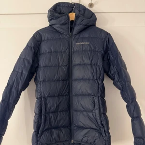 Peak Performance jacka  - Snygg och funktionell mörkblå dunjacka från Peak Performance med dragkedja och huva. Perfekt för kyliga dagar som håller värmen. 