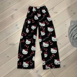 Svarta Hello Kitty byxor - Mysiga svarta byxor med Hello Kitty-mönster i vitt och rosa. Perfekta för en avslappnad dag hemma. Byxorna har en elastisk midja för extra komfort.