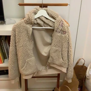 Beige teddyjacka med broderi - Mysig beige teddyjacka med broderad text 'Oh LA' och ett hjärta på bröstet. Jackan har en dragkedja framtill och långa ärmar. Perfekt för kyliga dagar när du vill vara både varm och stilren.