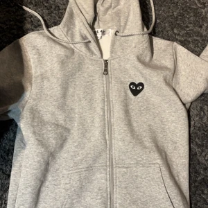 Grå hoodie från Comme des Garçons - Säljer en stilren grå hoodie från Comme des Garçons med dragkedja och det ikoniska hjärtmotivet med ögon på bröstet och ryggen. Perfekt för en avslappnad look.