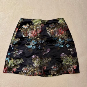 Svart blommig kjol - Elegant svart kjol med ett färgglatt broderat blommönster i rött, grönt, blått och guld. Kjolen har en kort längd och är perfekt för att ge en färgklick till din outfit. Materialet ser ut att vara glansigt (silk liknande) och ger en lyxig känsla.