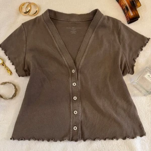 Brun kortärmad topp från Brandy Melville - Säljer en brun kortärmad topp från Brandy Melville. Toppen har en v-ringad design med knappar framtill och vågiga kanter vid ärmslut och nederkant. Perfekt för en avslappnad stil.