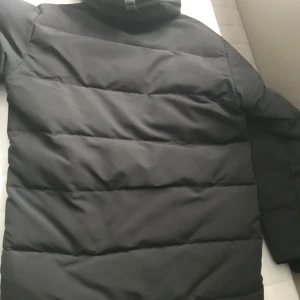 Svart pufferjacka från Canada Goose - Säljer en svart pufferjacka från Canada Goose med huva och dragkedja. Jackan har en broderad logotyp på ärmen och praktiska fickor framtill. Perfekt för kalla vinterdagar och den är m men passar L