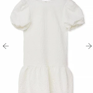 Vit klänning med puffärmar från NA-KD - Säljer en söt vit klänning från NA-KD med puffiga kortärmar och en fin knytning i ryggen. Klänningen har en volangdetalj vid fållen och är perfekt för sommarens alla tillfällen. Den är tillverkad i ett mönstrat tyg som ger en extra touch.