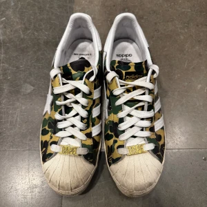 Adidas x Bape Camo Superstar - Unika Adidas sneakers i samarbete med Bape, med ett iögonfallande camouflagemönster i grönt, brunt och beige. Skorna har den klassiska Adidas-designen med vita ränder och snörning. Perfekta för dig som vill sticka ut! Storlek 41 1/3. 