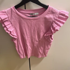 Rosa ribbad topp från Zara - Säljer en söt rosa ribbad topp från Zara med volangdetaljer på ärmarna. Perfekt för en somrig look. Toppen har en rund halsringning och är kortärmad. Lite skrynklig men går att fixa!!