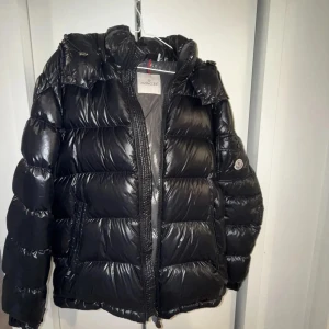 Moncler Maya - Säljer en snygg svart dunjacka från Moncler. Jackan har en glansig finish och är utrustad med dragkedja. Perfekt för kyliga dagar med sin varma och puffiga design. Storlek 2 som motsvarar s/m