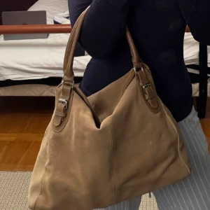 Beige axelväska i mocka - Snygg beige axelväska i mjukt mockamaterial med stilrena detaljer. Väskan har två handtag och spännen i metall som ger en elegant touch. Perfekt för att bära med sig det nödvändigaste med stil.