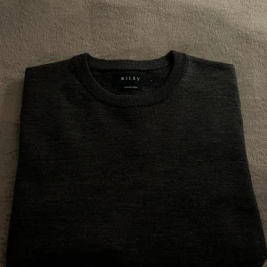 Mörkgrå tröja i merinoull från Riley - Säljer en stilren mörkgrå tröja från Riley i mjuk merinoull. Perfekt för kyliga dagar med sin klassiska design och bekväma passform. Ett måste i garderoben för den som älskar tidlös stil.