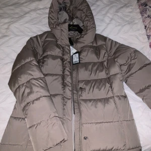 Beige dunjacka med huva - Snygg beige dunjacka från Lager 157 med huva och dragkedja. Perfekt för kyliga dagar med sin quiltade design som håller värmen. Jackan har långa ärmar och en stilren look. Aldrig använt❤️ 