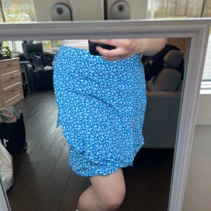 Blå blommig kjol från Bik Bok - Säljer en superfin blå kjol med vita blommor från Bik Bok. Kjolen har en omlott-design och knytband i sidan, vilket ger en söt volangdetalj. Perfekt för sommardagar eller en mysig vårutflykt. Den är i nytt skick med taggar kvar. 🌸