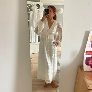 Vit långklänning med knytband - Säljer en superfin vit långklänning med v-ringning och knytband i midjan. Klänningen har långa ärmar och ett diskret rutmönster som ger en elegant touch. Perfekt för sommarens alla fester eller en mysig midsommar. Materialet känns lätt och luftigt.