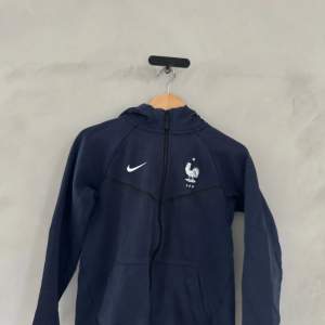 Säljer en snygg mörkblå hoodie från Nike med dragkedja och det franska fotbollsförbundets logga på bröstet. Perfekt för sportiga tillfällen eller bara för att chilla. Den har långa ärmar och en bekväm passform. Passar perfekt till höst och vår!