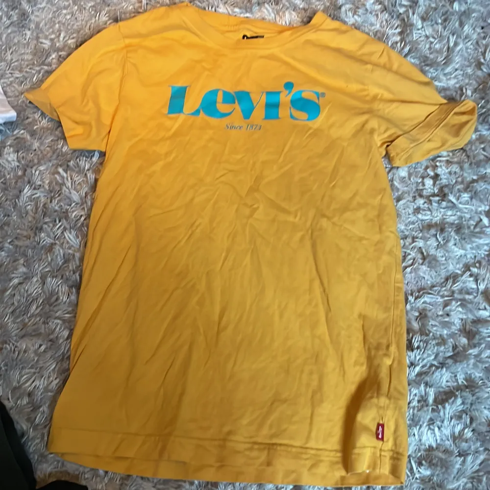 Säljer en snygg gul t-shirt från Levi's i bomull. Den har en stor blå logga på bröstet och är perfekt för sommardagar. T-shirten är kortärmad och i bra skick. Perfekt för en avslappnad stil!. T-shirts.