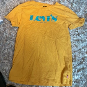 Gul t-shirt från Levi's - Säljer en snygg gul t-shirt från Levi's i bomull. Den har en stor blå logga på bröstet och är perfekt för sommardagar. T-shirten är kortärmad och i bra skick. Perfekt för en avslappnad stil!