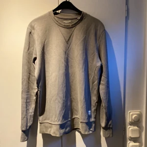 Grågrön sweatshirt från Jack & Jones - Säljer en stilren grågrön sweatshirt i bomull från Jack & Jones Essentials i storlek L. Tröjan har en klassisk rund hals och långa ärmar. Passar bra till jeans eller chinos för en avslappnad look.