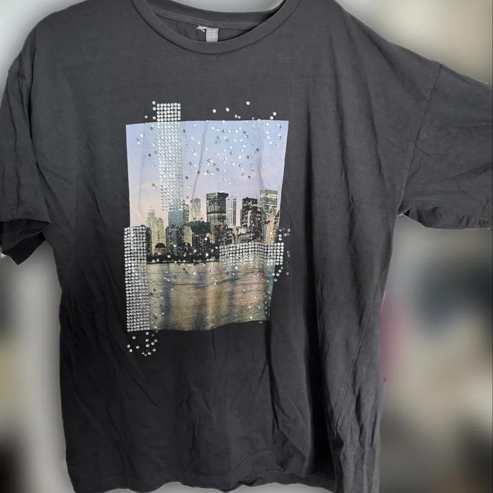 Säljer en mörkgrå t-shirt med en cool stadsbild på framsidan. Storlek 42/L. Den har strassdetaljer som ger en glittrig effekt över motivet. Knappt använd och inga synliga defekter. . T-shirts.