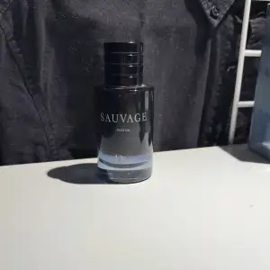 Säljer en flaska Sauvage Parfum från Dior. Flaskan är elegant och mörk med en stilren design. Den har en svart kork med CD-loggan på toppen. Perfekt för den som vill ha en lyxig och maskulin doft. Passar både till vardags och fest. Flaskan är i gott skick och redo att användas. (OBS) parfymen är använd alltså inte helt full.