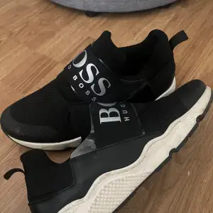 Snygga svarta sneakers från Hugo Boss i mycket bra skick. De har en elastisk rem med logotyp över foten och en bekväm vit sula. Perfekta för både vardag och träning. Skorna har en modern design med en stilren look.