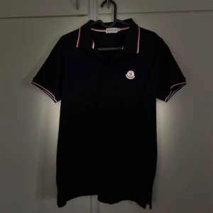Svart moncler piketshirt  - Säljer en snygg svart pikétröja från Moncler. Den har korta ärmar och en klassisk krage med röd och vit rand. Perfekt för både vardag och fest! Tröjan har en broderad logga på bröstet som ger en exklusiv touch.