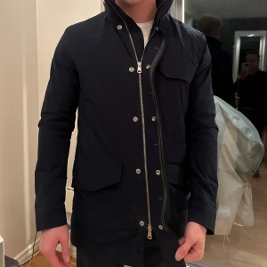 J.Lindeberg jacka - Längre mörkblå jacka från J.Lindeberg i strl S. Mycket bra skick, använd sparsamt 1 vinter. Lite skrap på märket som sitter på vänster arm, annars inga defekter.