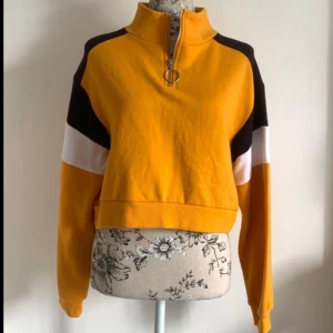 Gul och svart croppad tröja med dragkedja - Säljer en supercool gul tröja med svarta och vita detaljer. Den har en halv dragkedja med en ringdetalj och är croppad för en trendig look. Perfekt för höst och vår! Den är långärmad och gjord i ett mjukt material som känns skönt mot huden.