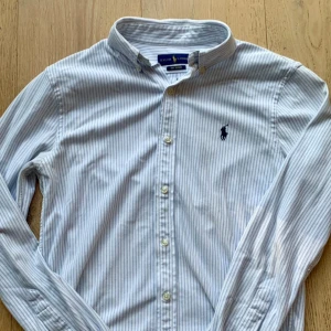 Blåvit randig skjorta från Ralph Lauren - Säljer en snygg och klassisk blåvit randig skjorta från Ralph Lauren i toppenskick. Skjortan är mycket stilren och har en broderad Ralph Lauren logga på bröstet.