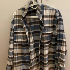 Rutig skjorta från H&M - Säljer en rutig skjortajaxcka från H&M. Den har en klassisk krage och knappar framtill. Skjortan är i en mix av blå, brun och beige färger, perfekt för höst och vår. Mycket bra skick och knappt använd 