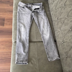 Grå jeans från Jack & Jones - Säljer ett par snygga grå jeans från Jack & Jones i modellen Slim/Glenn. De har en slim passform och är tillverkade i ett bekvämt jeansmaterial. Byxorna har knappar i gylfen och klassiska fem fickor. Perfekta för en casual look!