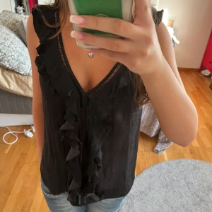 Svart ärmlös topp med volangdetaljer - Säljer en snygg svart ärmlös topp från H&M. Den har en v-ringad design med fina volangdetaljer längs framsidan. Perfekt för både vardag och fest!