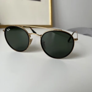 Ray-Ban Round Double Bridge - Säljer ett par UV-skyddade runda Ray-Ban storlek 51 med svart glas och gulfärgad bågar. Helt nya, använt 1ggn  därmed nyskick! Nypris:1500kr  Mått: Näsbrygga:22mm Glasbredd:51mm Skalmlängd:145mm 
