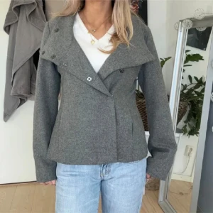 Grå ulljacka med hög krage - Säljer en snygg grå ulljacka med hög krage och dubbelknäppning. Jackan har en stilren design med långa ärmar och en skön passform. Perfekt för höst och vår när det är lite kyligare ute. Den är i bra skick och passar till både jeans och mer uppklädda outfits.