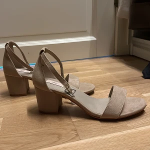 Beige klackskor från Nelly - Säljer ett par snygga beige klackskor från Nelly. De har en bred rem över tårna och en justerbar rem runt ankeln. Skorna har en stabil blockklack som gör dem bekväma att gå i. Perfekta för både vardag och fest! ALDRIG ANVÄNDA!!!