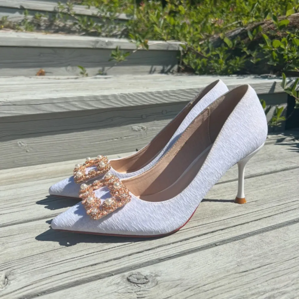 Säljer ett par eleganta vita pumps i ett lite skimmrigt tyg, med spetsig tå och smal klack. Skorna har en vacker dekoration framtill med pärlor och glittrande stenar, liknar manolo blahnik. Perfekta för fest, i storlek 40 men närmare storlek 39. . Skor.