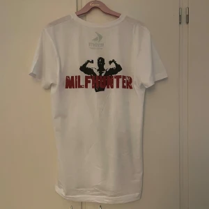 Revenite Milf hunter t shirt  - Säljer en vit t-shirt från Revenite i storlek S. Den har ett tryck på baksidan med texten 'MILF HUNTER' och en bild av en person som flexar musklerna. Perfekt för en avslappnad stil eller som statement-plagg. Tillverkad i Sverige. Går ej att få tag i