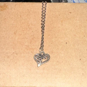 Silverfärgat halsband med hjärthänge - Säljer ett fint silverfärgat halsband med ett detaljerat hjärthänge. Hänget har ett intrikat mönster som ger en elegant touch. Perfekt för både vardag och fest!