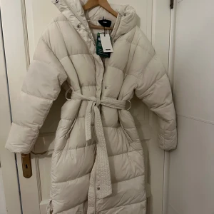 Vit lång pufferjacka från Bik Bok - Säljer en supermysig och varm vit pufferjacka från Bik Bok. Den är lång och perfekt för kalla vinterdagar. Jackan har ett bälte i midjan för en snygg passform. Den är i nyskick och har både dragkedja och knappar. Aldrig använd. 